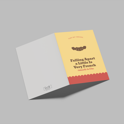 A folded greeting card shown open on a plain gray background, with the front cover in pale yellow, a red scalloped band, and the back cover in white. The front reads 'Falling Apart a Little Is Very French' with a small croissant illustration.